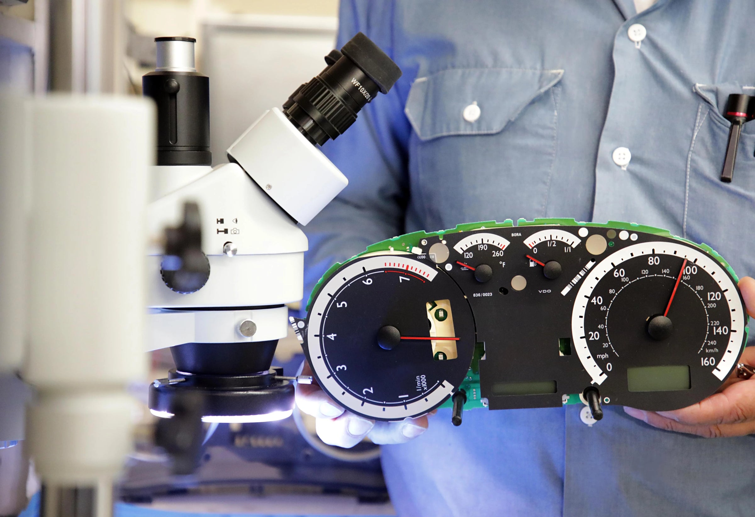 Technician holding a disassembled automotive instrument cluster in front of a microscope during electronic repair inspection.